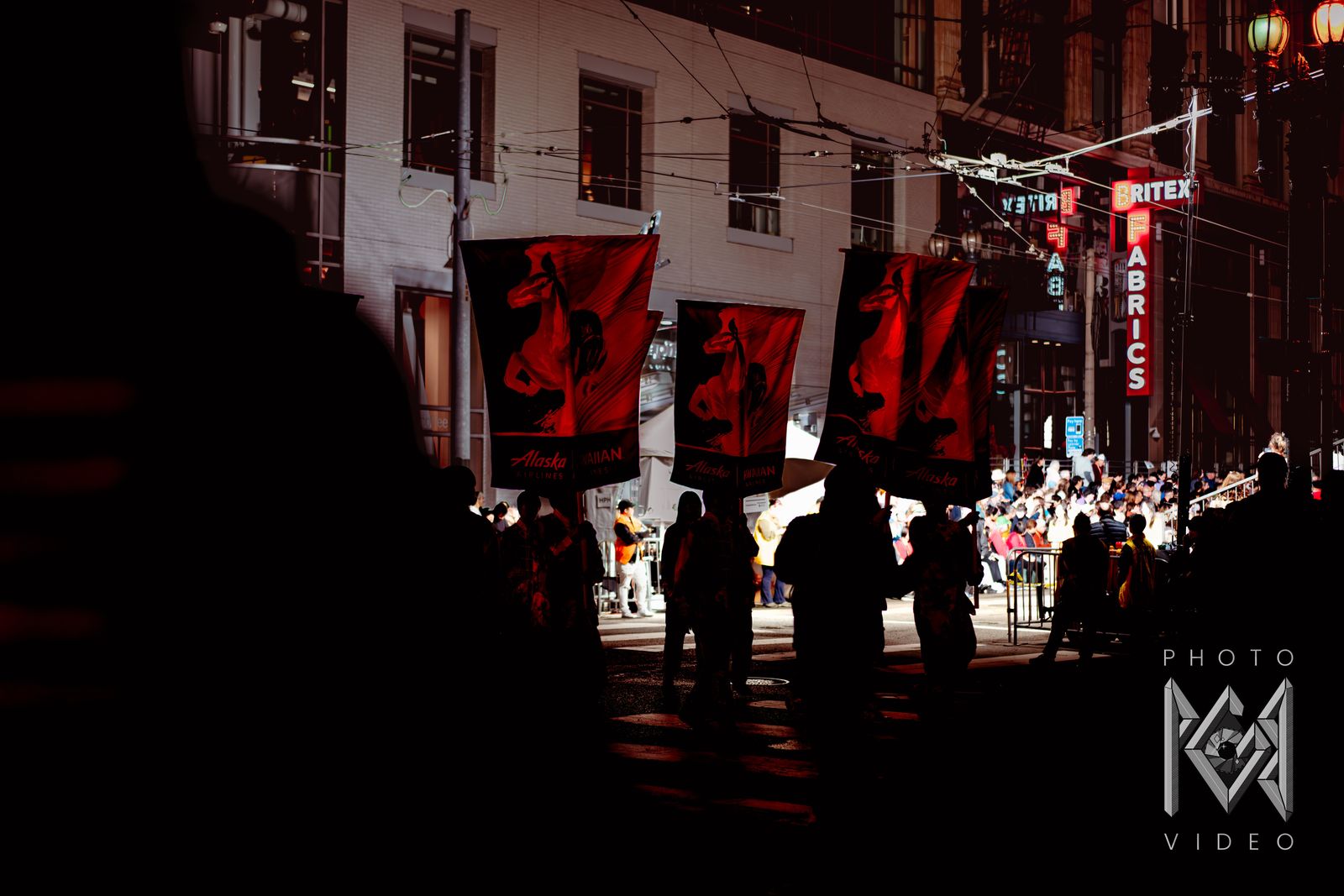 Parade marchers in silhouette carrying red horse banners past the glowing Britex Fabrics sign