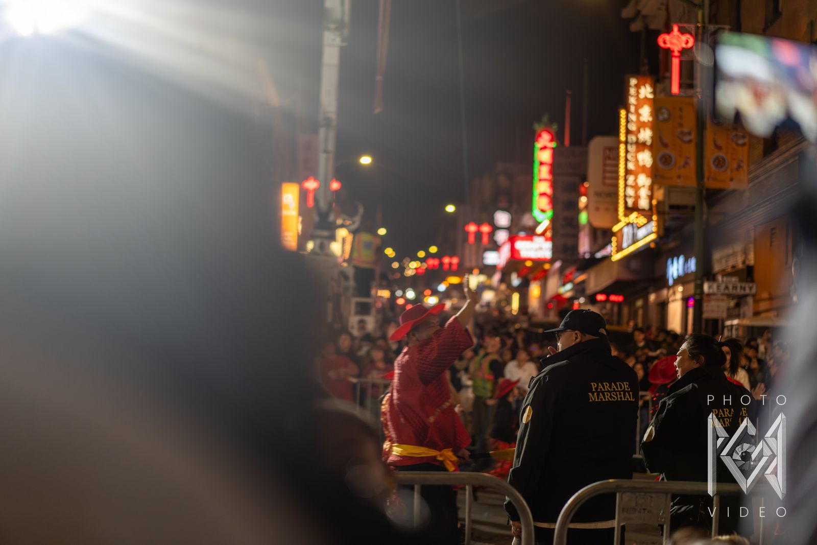 A performer in a red hat waves to the crowd as parade marshals look on beneath Chinatown's neon signs
