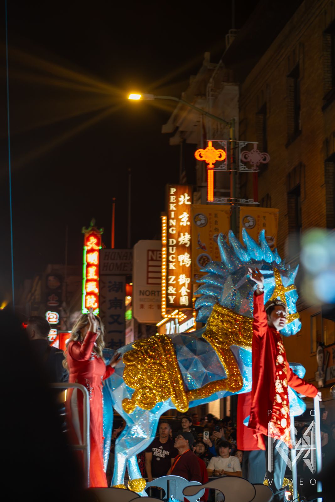 Performers in red wave from a glittering blue and gold horse float beneath the Peking Duck neon sign