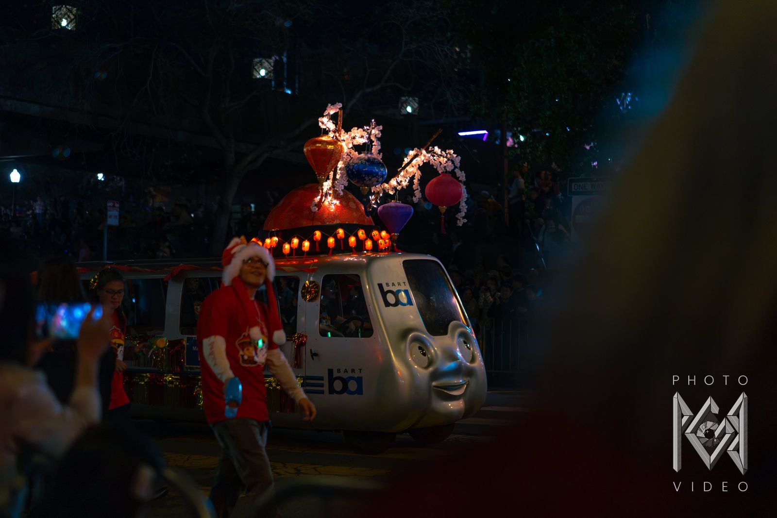 A smiling BART train float topped with lanterns and blossom branches rolls past the crowd