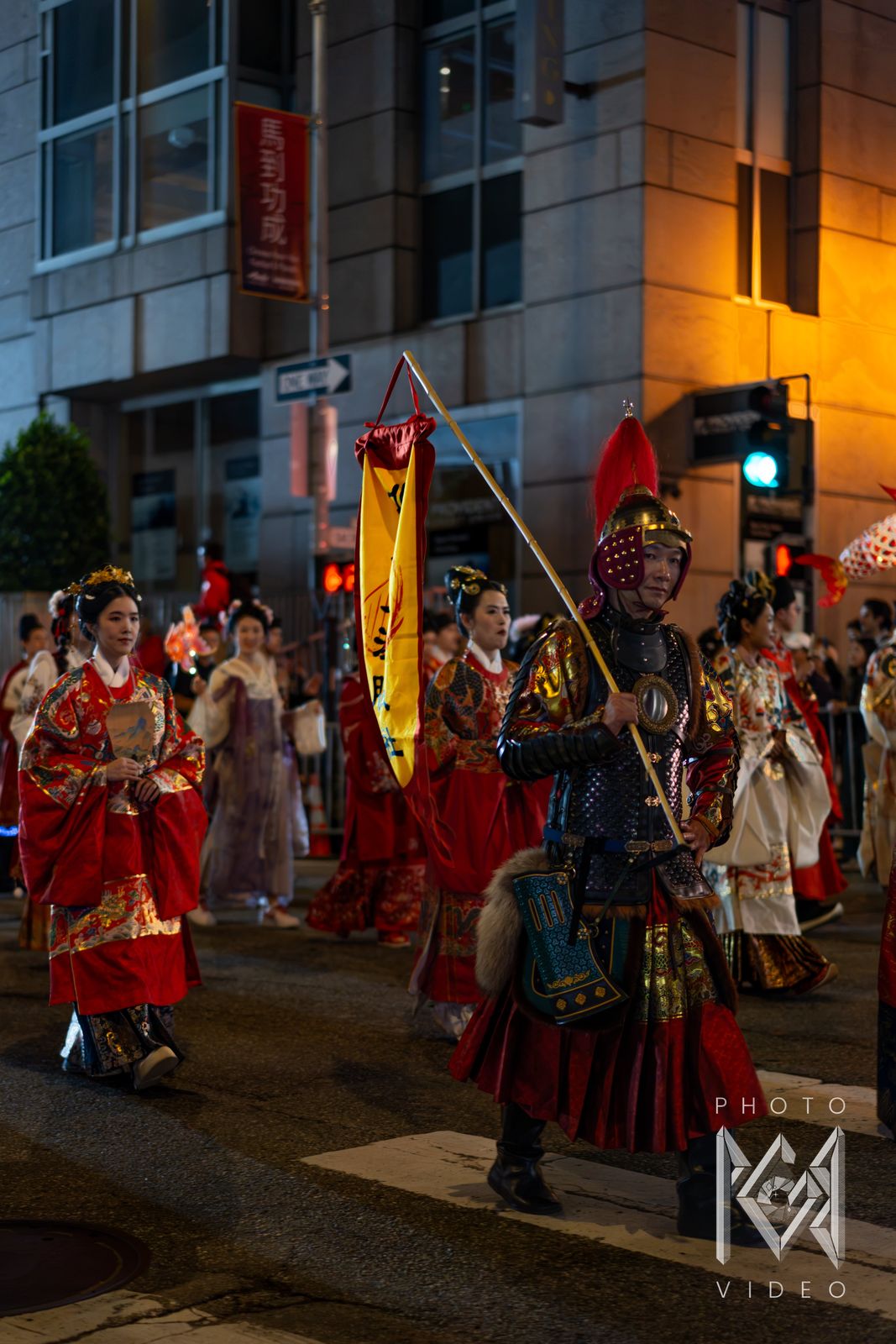 A warrior in armour and a red-plumed helmet carries a yellow banner, followed by women in red embroidered robes