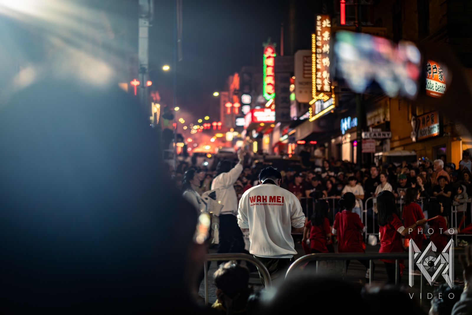 Crowds lining a Chinatown street lit by neon signs, with a bright floodlight overhead
