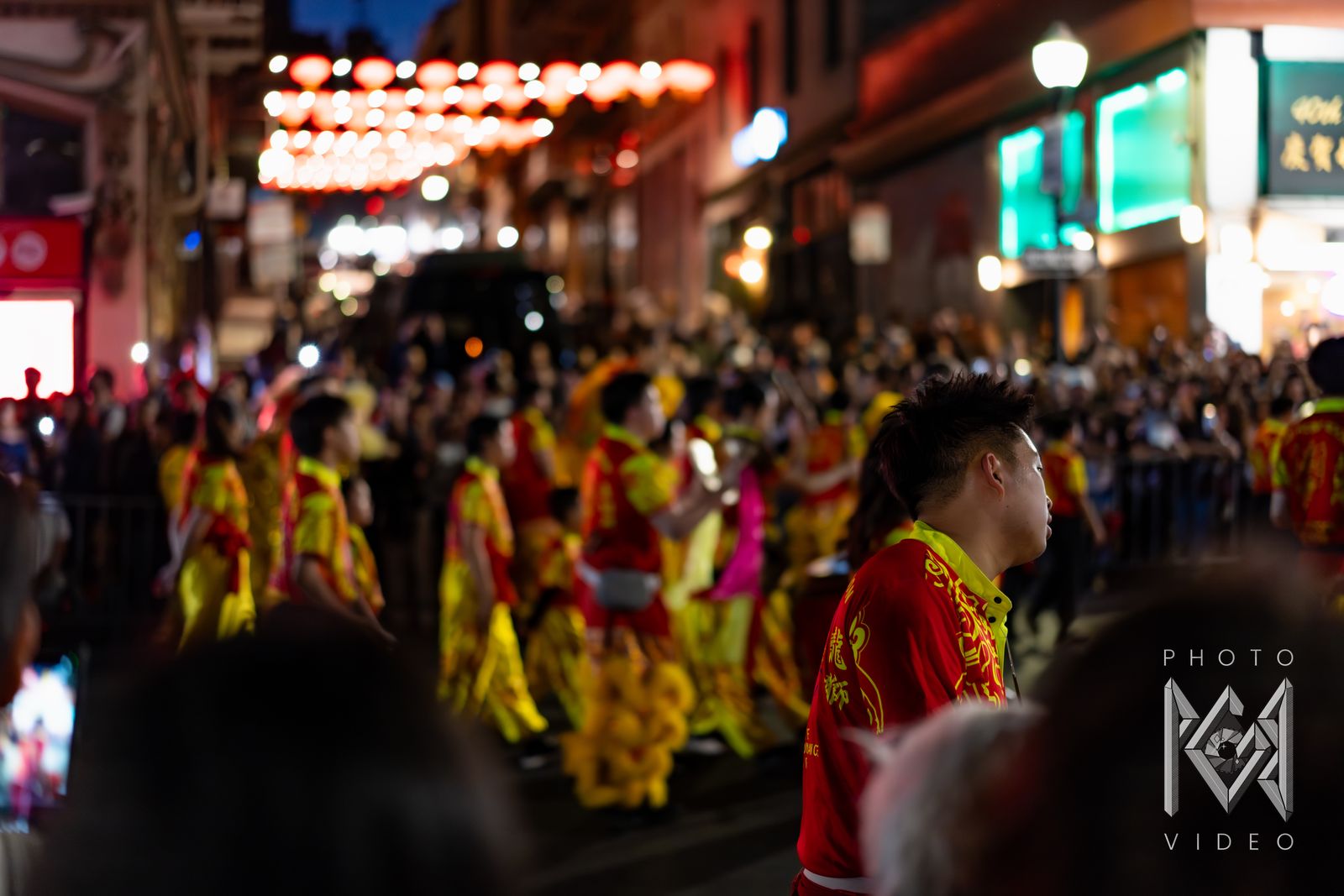 Dancers in red and yellow costumes moving through a packed street under strings of red lanterns