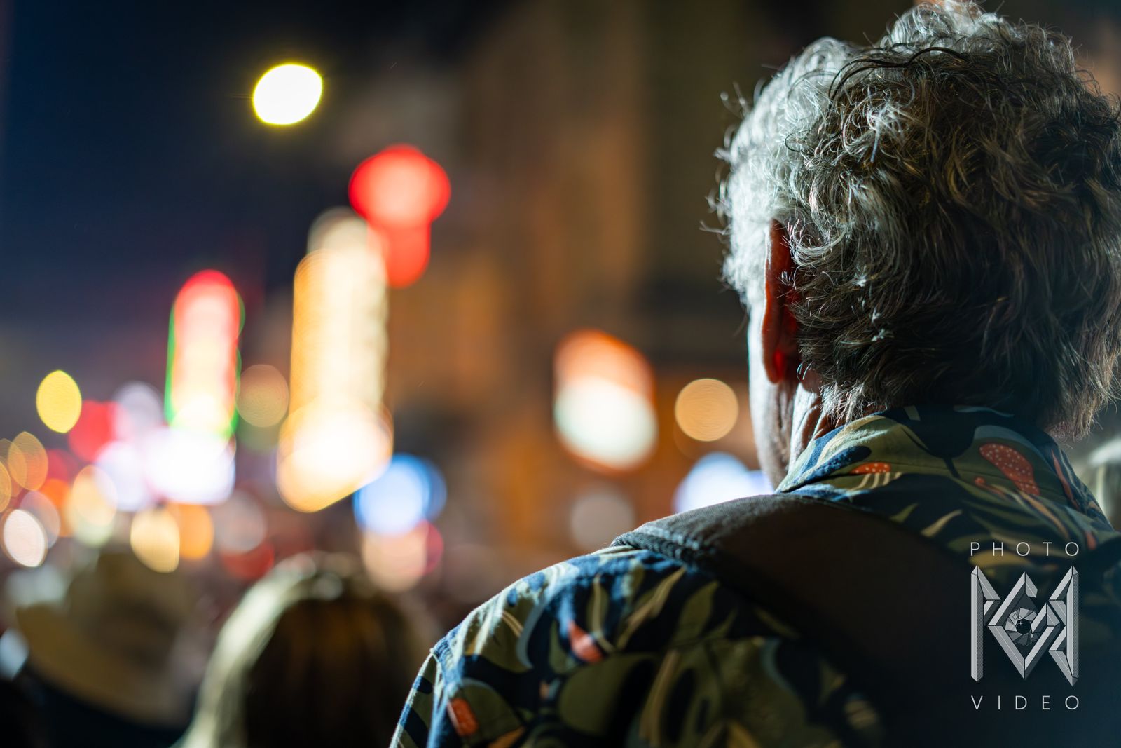 A grey-haired spectator in a patterned shirt watching the parade, Chinatown&rsquo;s neon blurred into soft circles of light