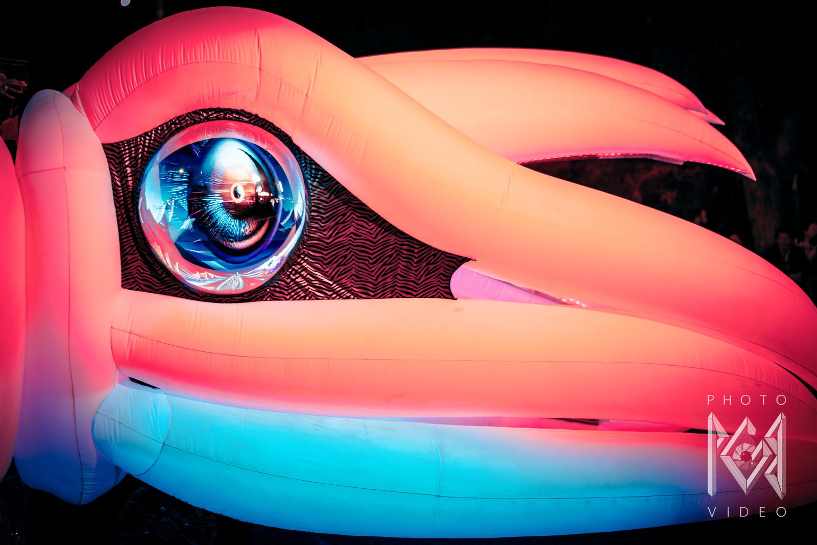 Close-up of a giant inflatable bird head glowing pink and blue, with a mirrored eye