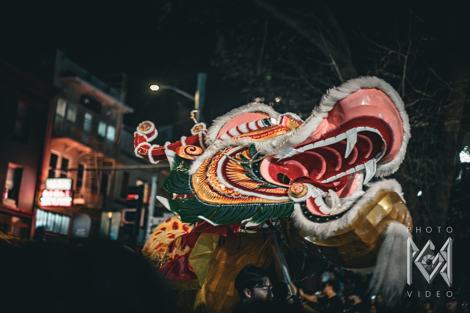 A dragon dance head with an open fanged mouth rearing above the crowd on a Chinatown street