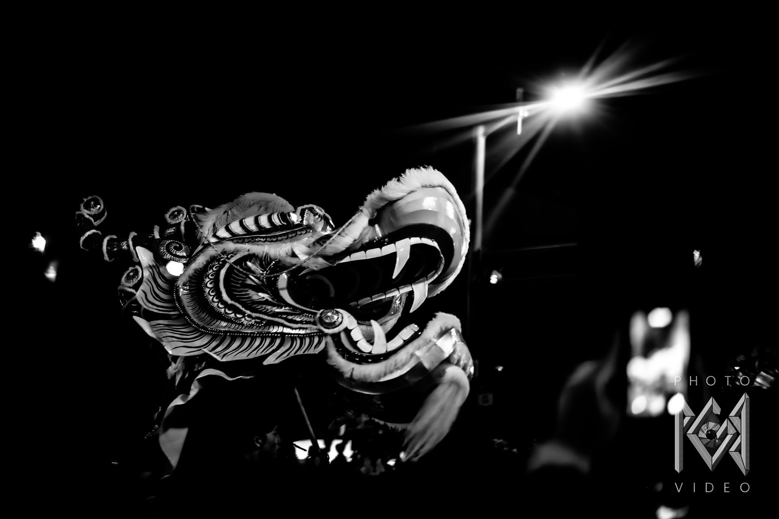 A dragon dance head with an open fanged mouth lit by a street lamp, in black and white