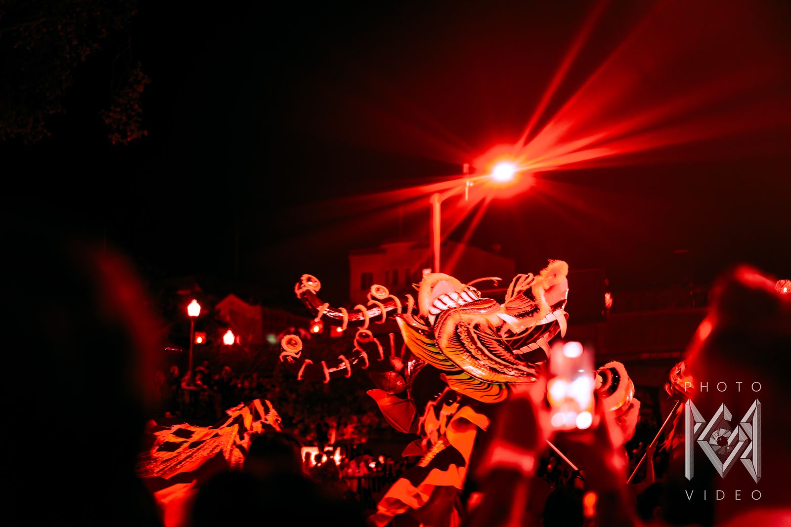 A dragon dance winding through a crowd of raised phones, all bathed in red light under a glaring street lamp