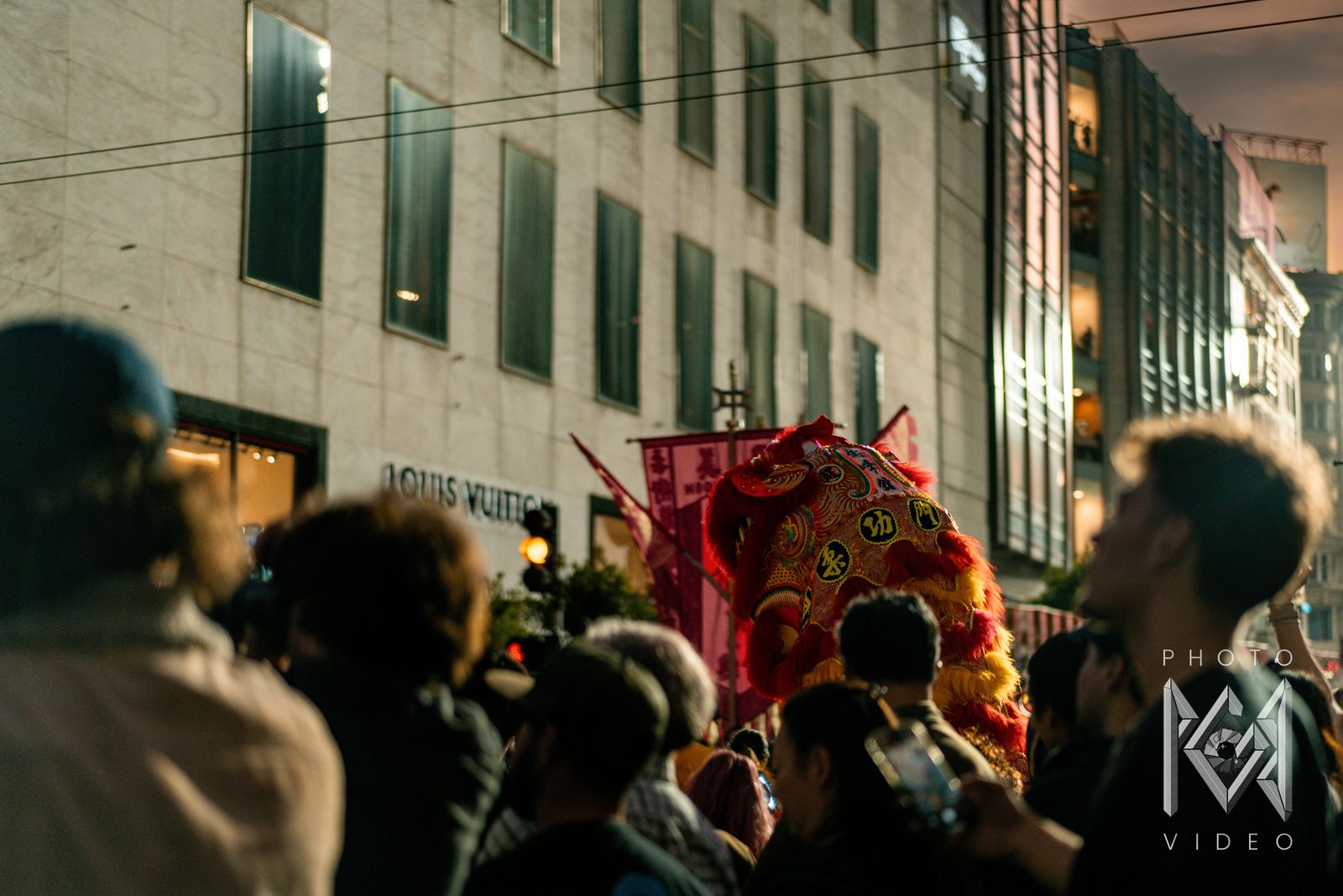 A red and gold lion dance head rising above the crowd in front of the Louis Vuitton store, with pink banners behind