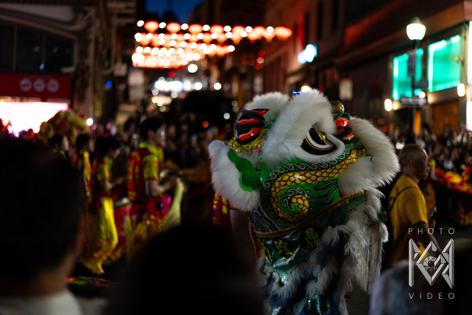 A green and white lion dance head turning through the crowd under strings of red lanterns