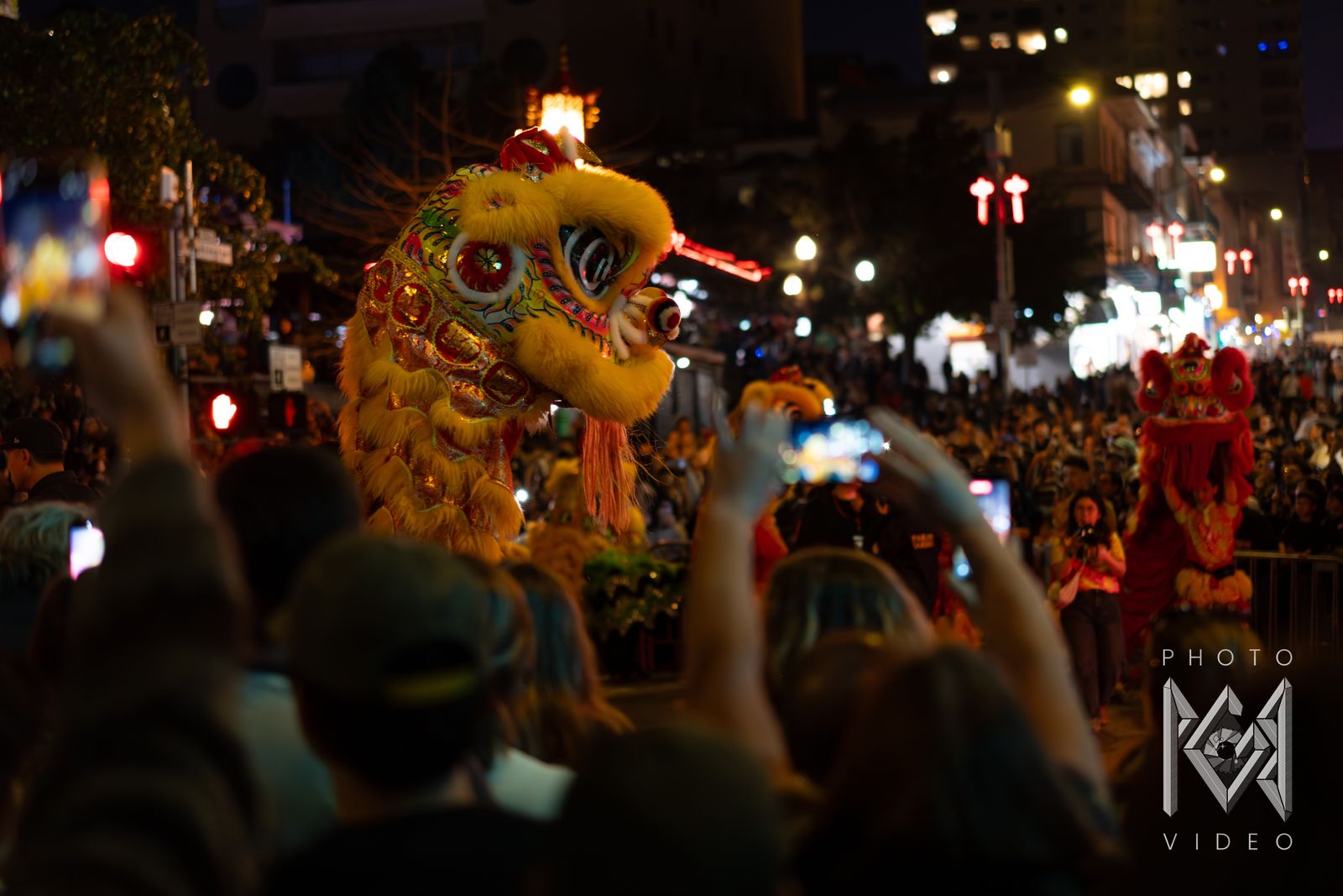 A yellow lion dance head rearing over a crowd of raised phones, with a red lion further down the street