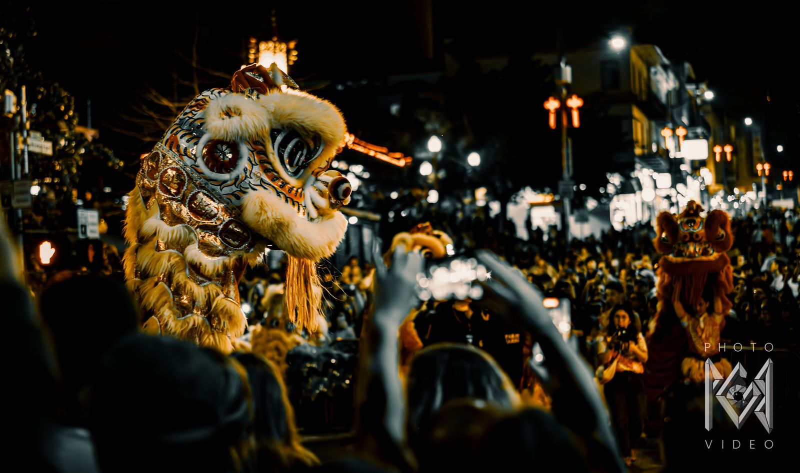 A yellow lion dance head rearing over a crowd of raised phones, in muted teal and gold tones