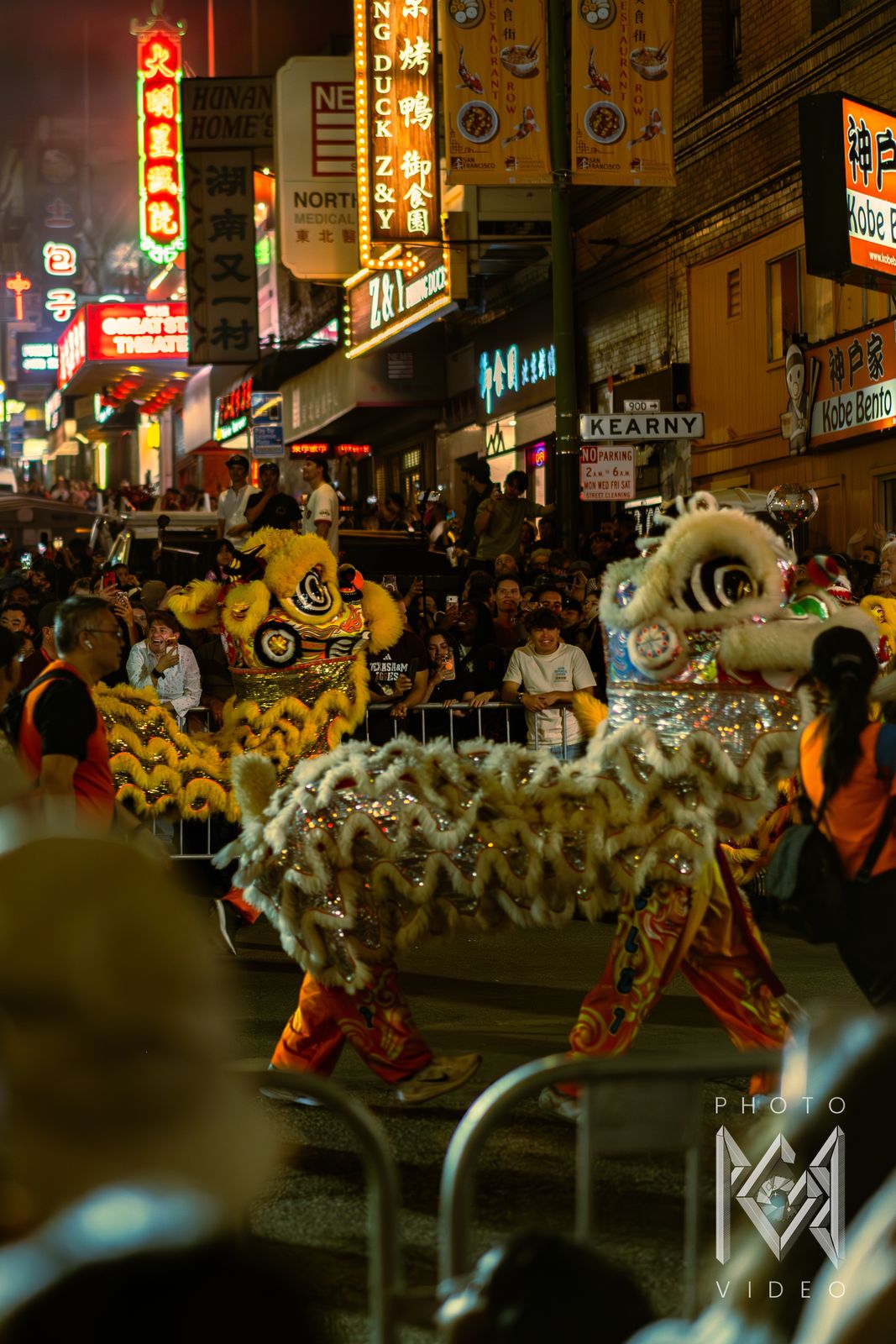 A white lion and a yellow lion dancing past spectators along Kearny Street beneath glowing Chinatown signs