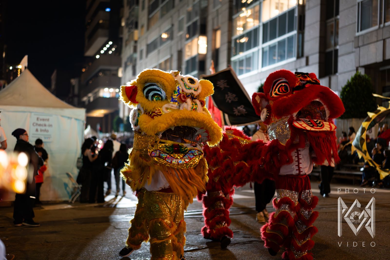 A yellow lion and a red lion dancing side by side down the street beside an event tent