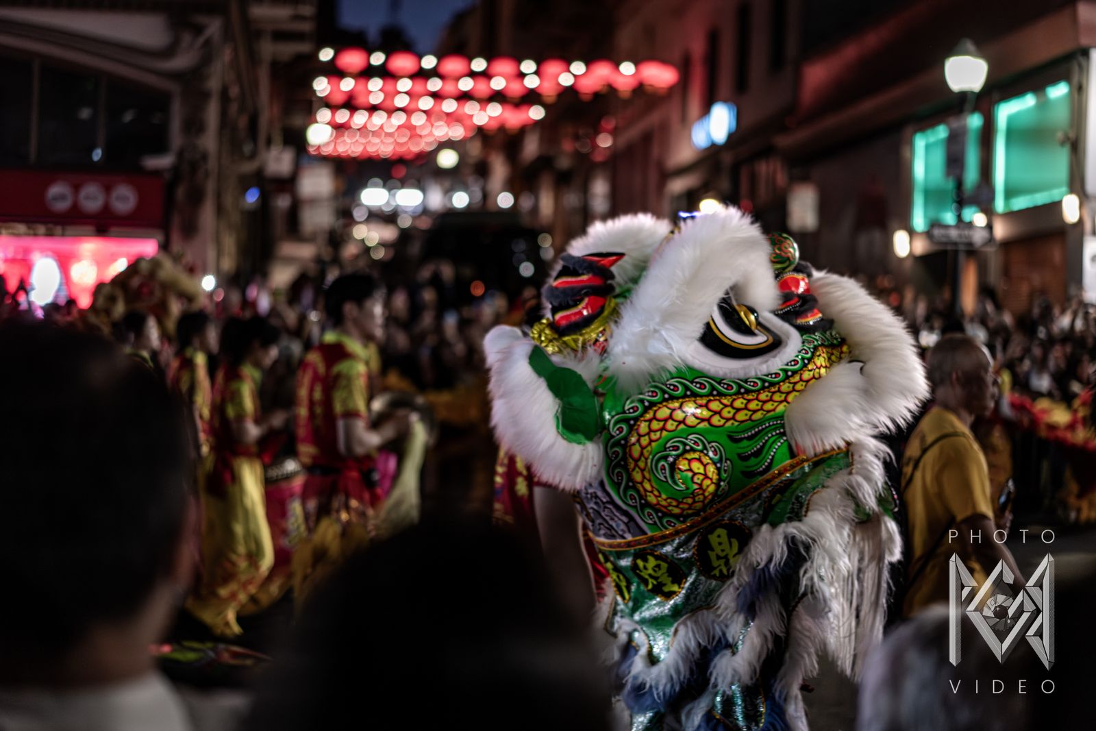 A green and white lion dance head turning through the crowd under red lanterns, in darker, cooler tones