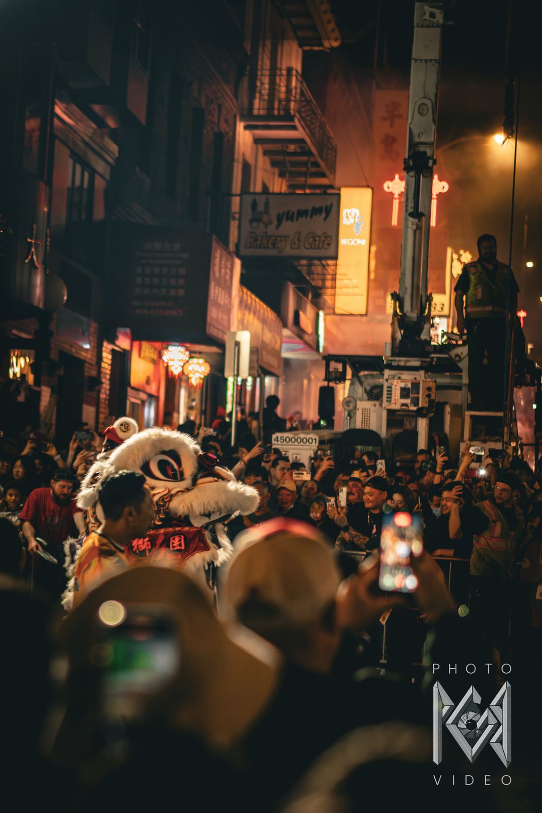 A white lion dancer moving through a packed crowd below the Yummy Bakery & Cafe sign