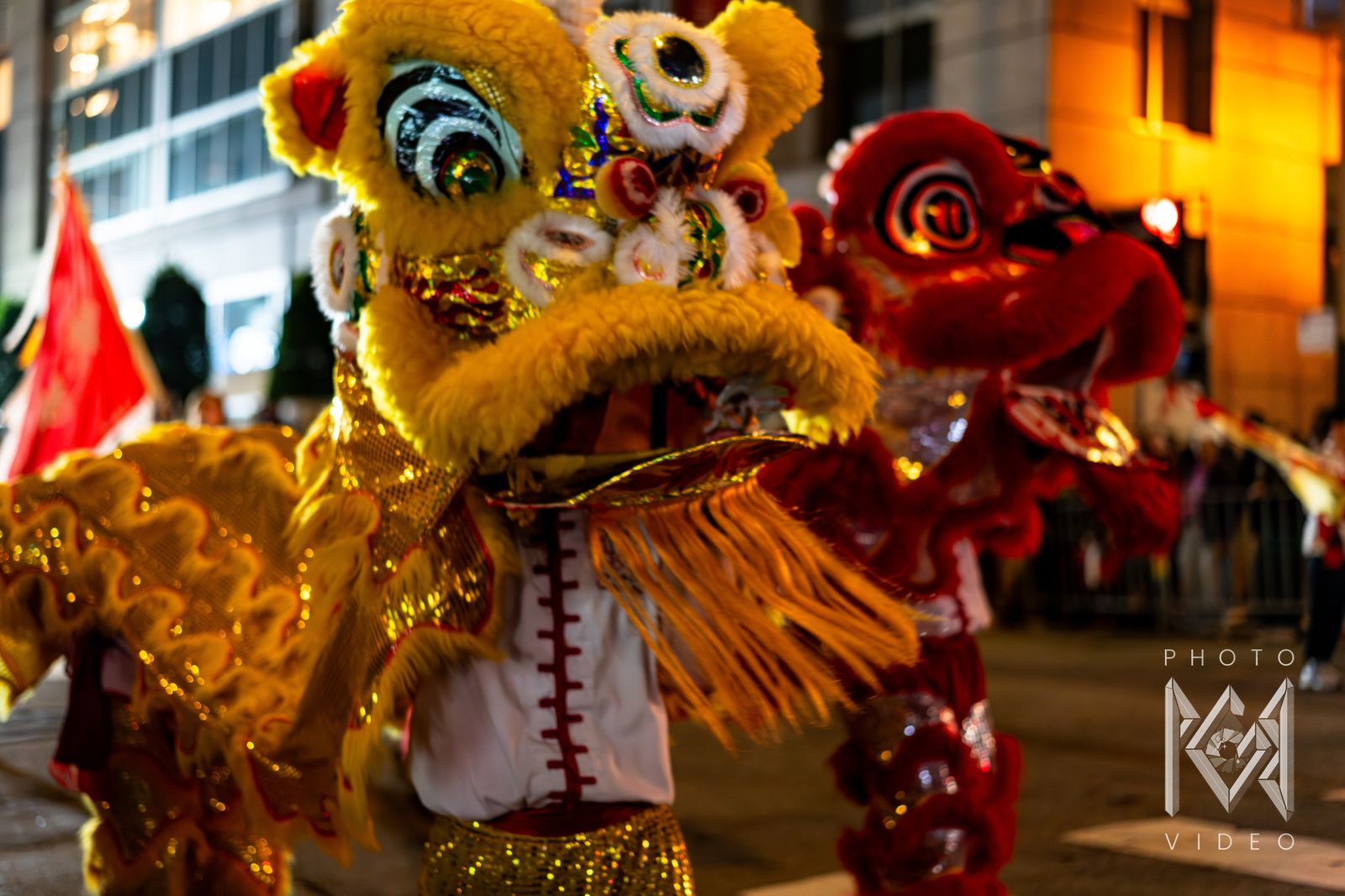 A yellow lion dance head in close-up with a red lion dancing just behind it on a lit street