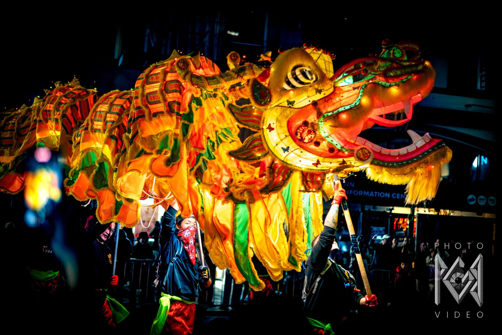 A glowing yellow and orange dragon head with an open mouth, carried on poles by dancers in the dark