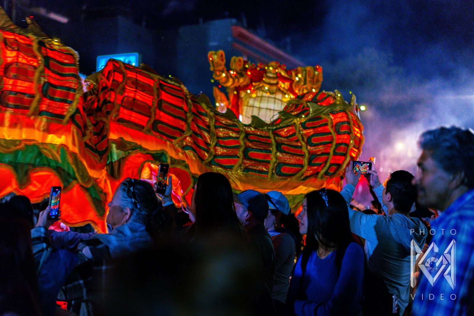 Spectators holding up phones beneath the glowing red and gold body of a dragon in a misty street