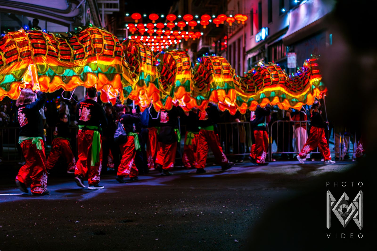 A long glowing dragon carried along a street under strings of red lanterns by dancers in red trousers