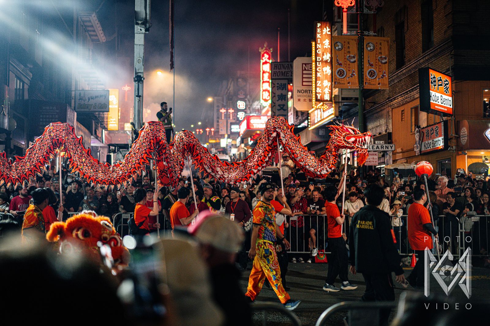 A long red dragon held high on poles above Kearny Street, with crowds behind the barriers and neon signs beyond