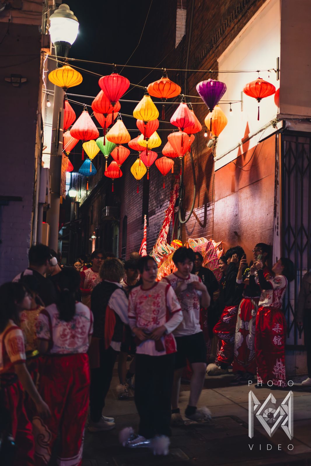 Dancers in red and white costumes resting in a Chinatown alley beneath strings of coloured silk lanterns