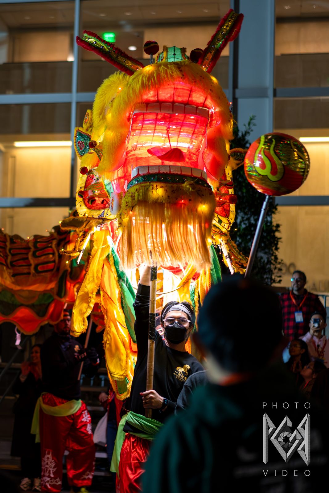 A glowing yellow dragon head with an open red mouth raised on a pole by a masked dancer, with the pearl beside it