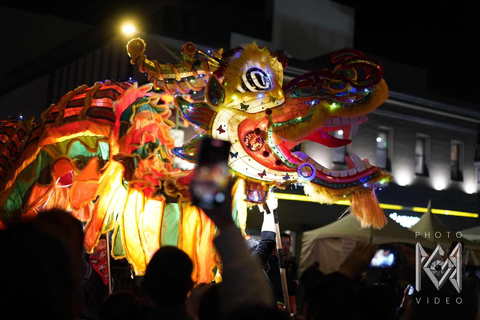 A glowing yellow dragon head with an open mouth and fangs, lit with fairy lights, as spectators raise their phones