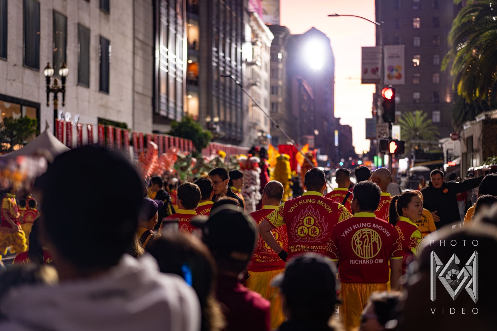 Kung fu school members in Yau Kung Moon jerseys walking with the parade toward a pink sunset sky