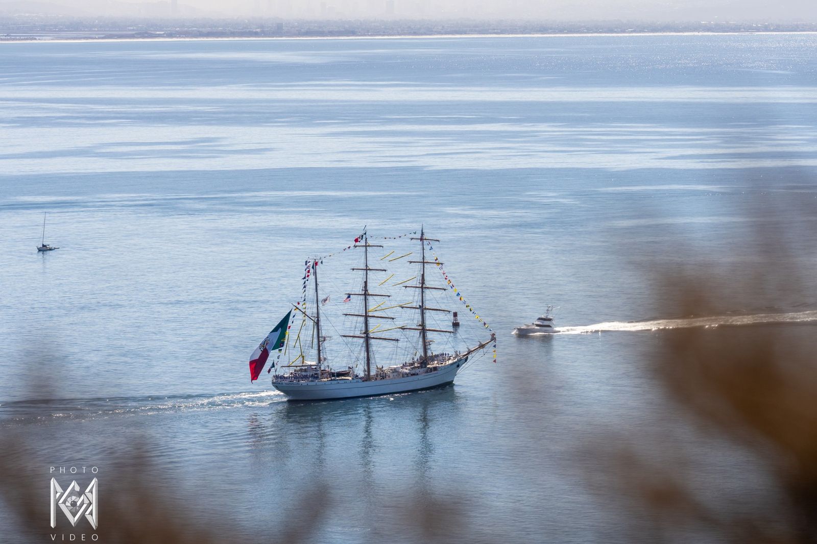 The tall ship under way on calm water with a Mexican flag at her stern and a small boat in her wake