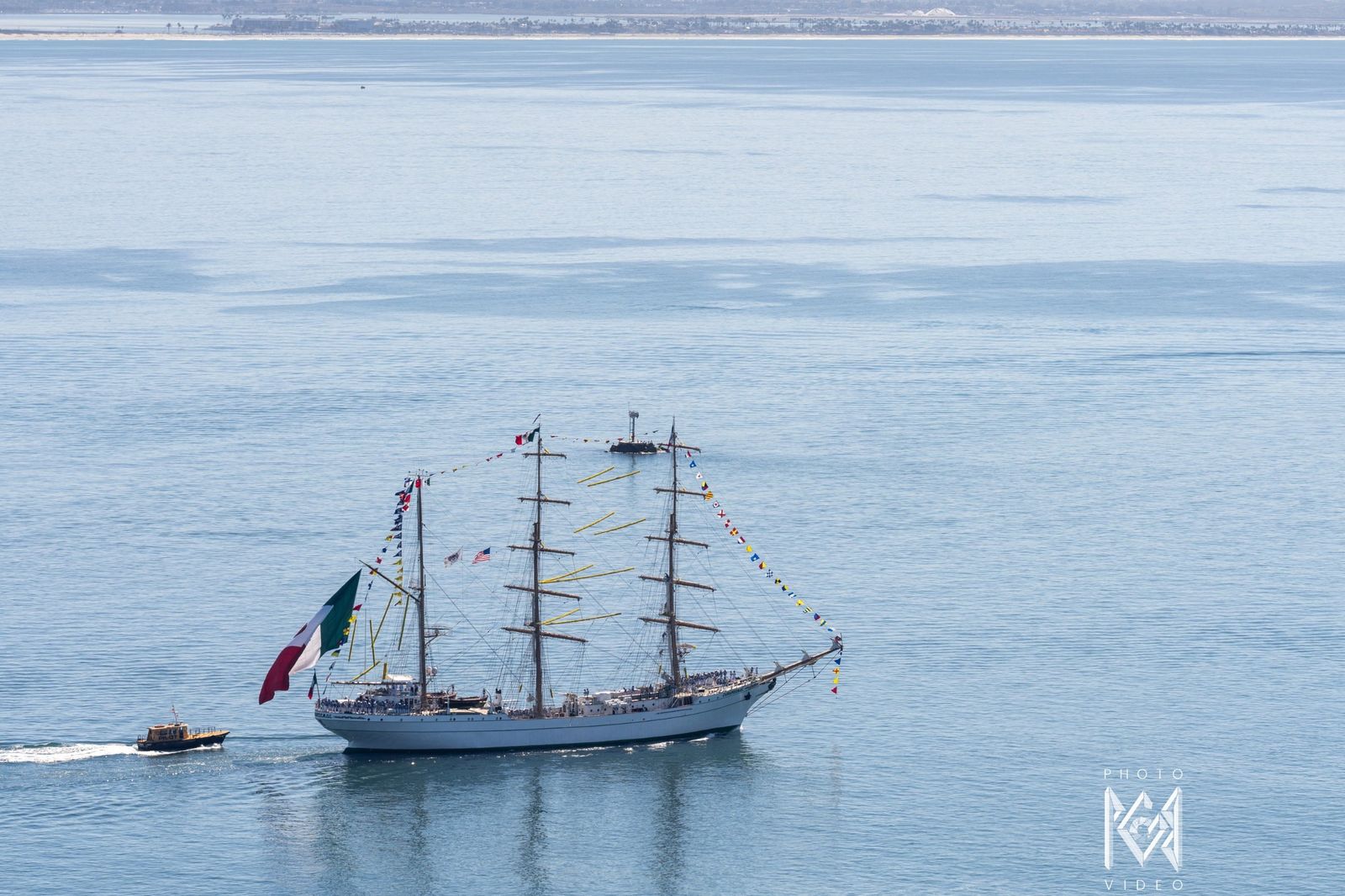 The tall ship moving across smooth blue water with a Mexican flag at the stern and a small boat alongside
