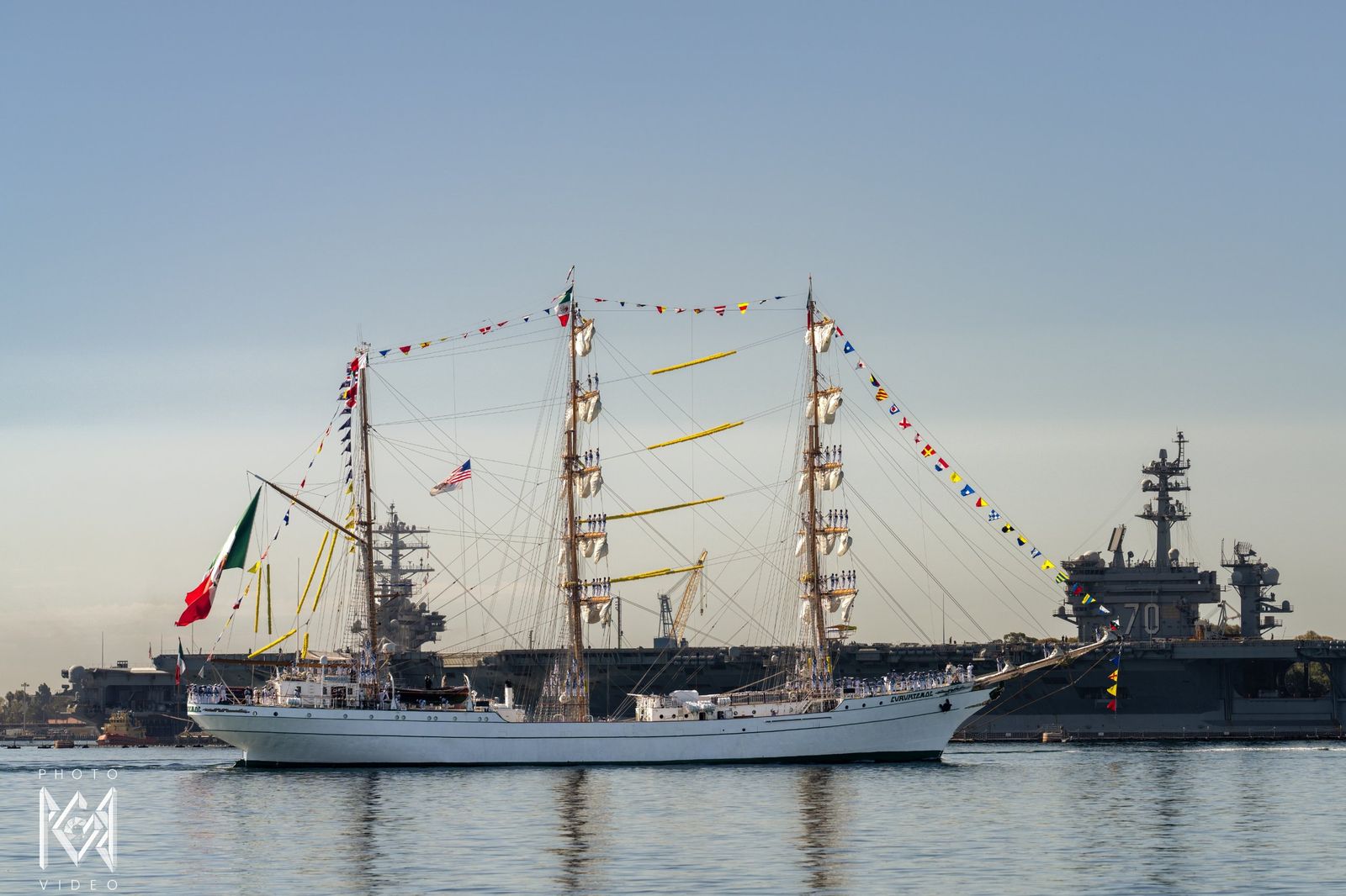The tall ship dressed with flags passing a grey aircraft carrier at the naval base