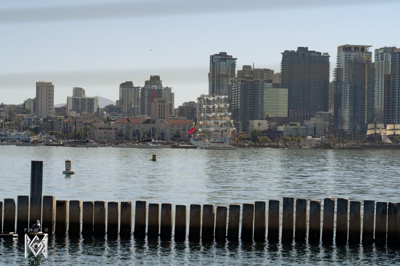 Distant city towers across the water with the ship's masts among them, behind a row of pier posts