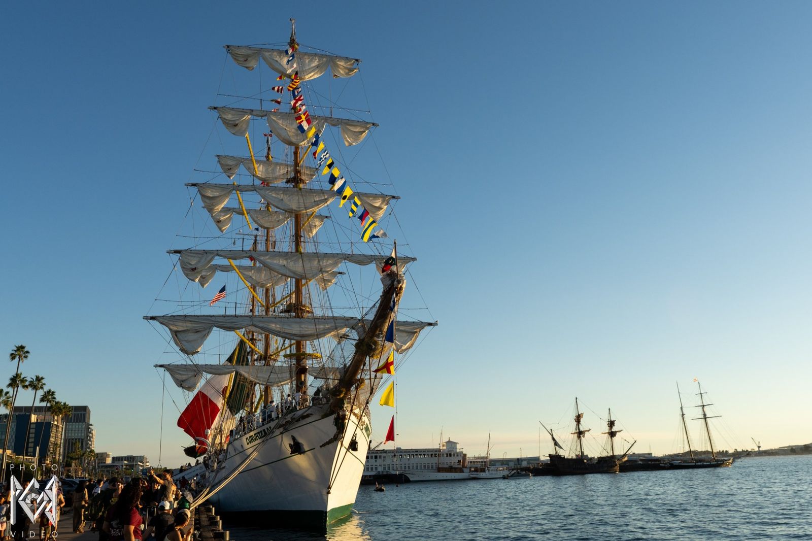 The ship moored at the pier at dusk, a crowd along the waterfront and sailboats beyond