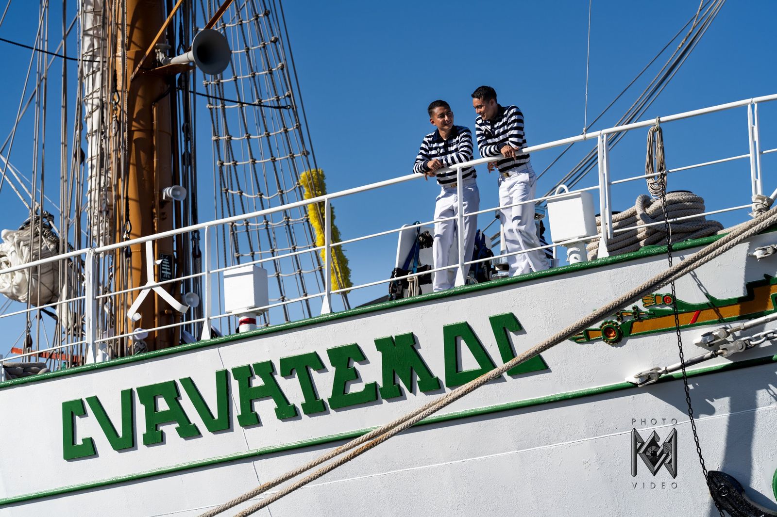 Two crew members in navy and white standing at the ship's rail above her green lettering