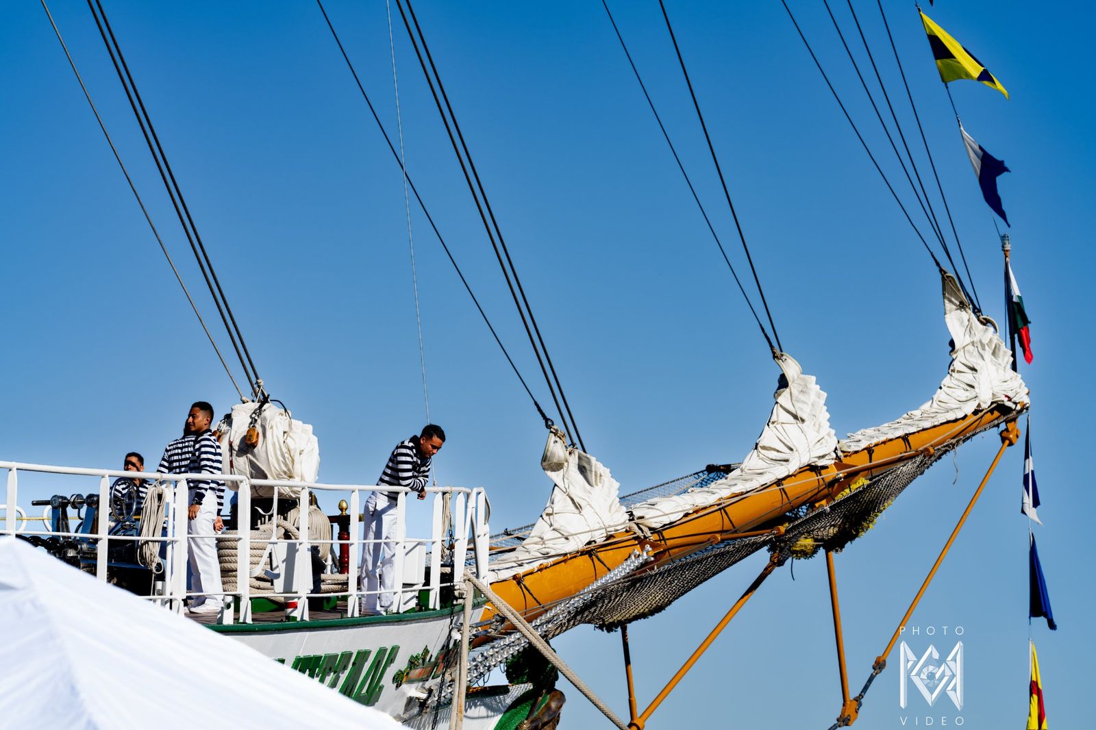 Crew standing at the rail beside the bowsprit under a blue sky, flags strung above