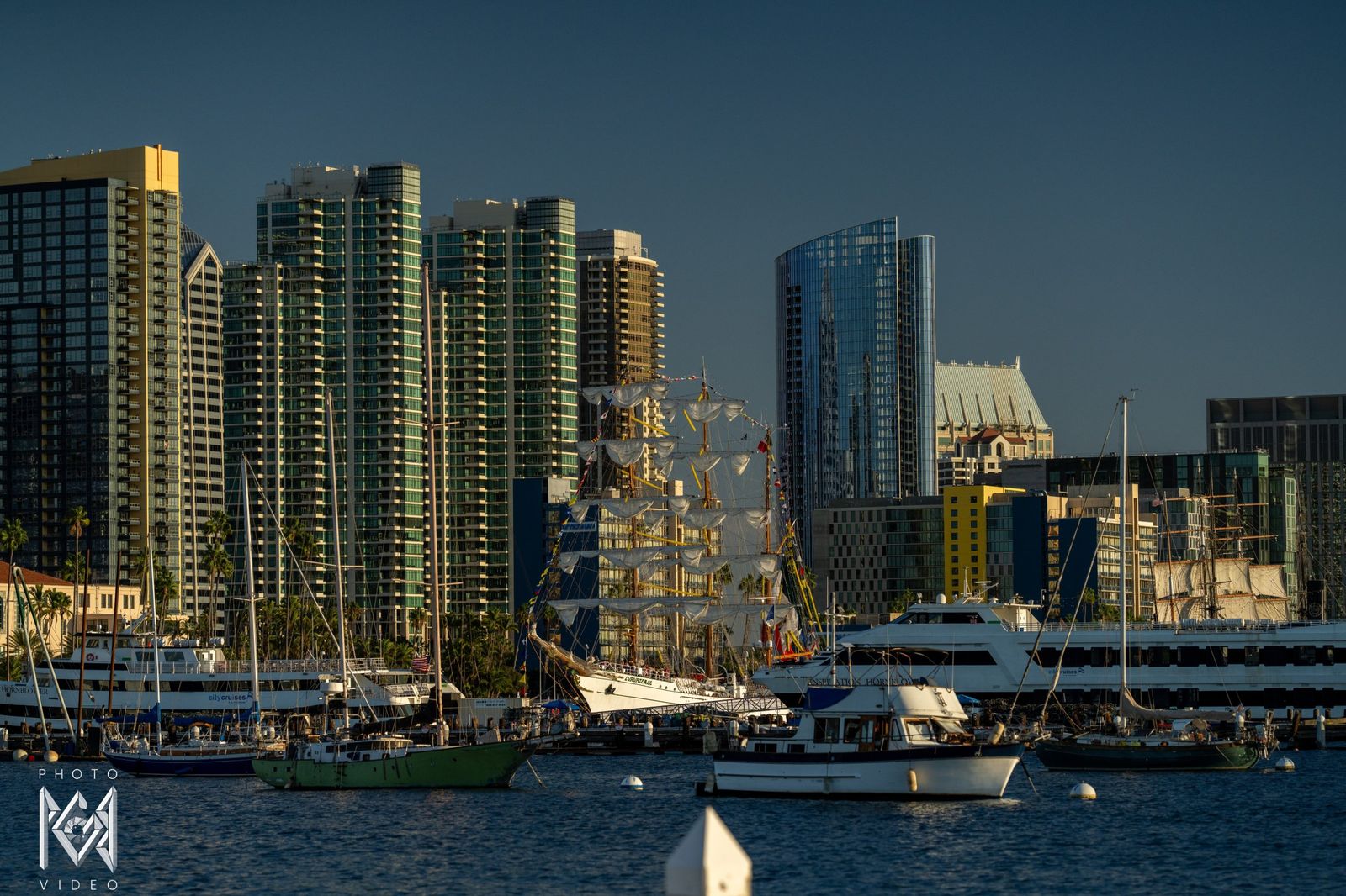 City towers across the harbour with boats moored in front and the ship's masts behind