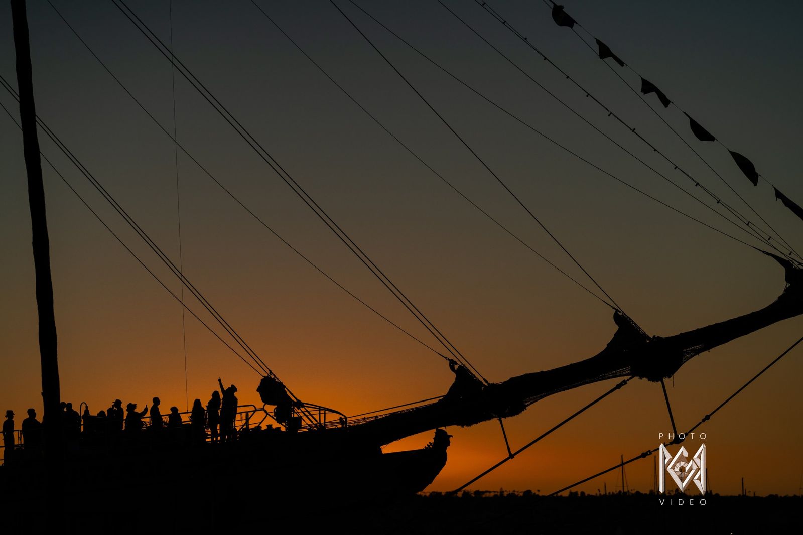 The bowsprit and a line of crew in silhouette against an orange sunset sky