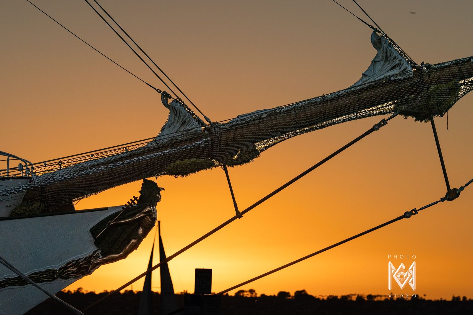 The bowsprit and stays in silhouette against a deep orange sky