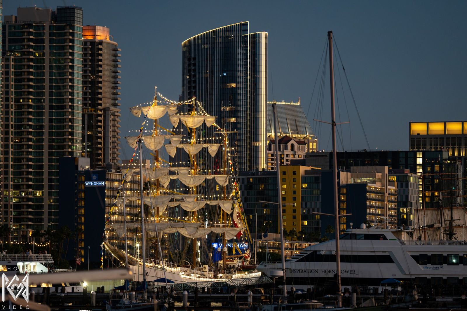 The ship lit among city towers at dusk, boats moored in front