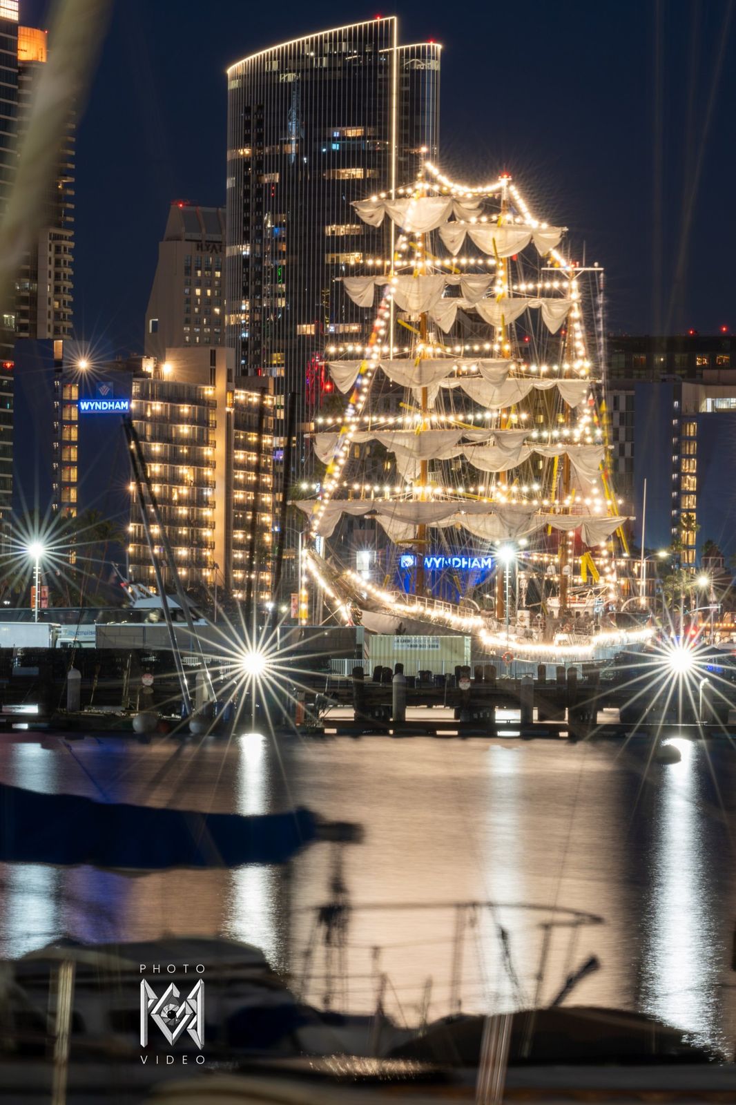 The ship outlined in lights at night in front of a lit tower, its reflection in the harbour