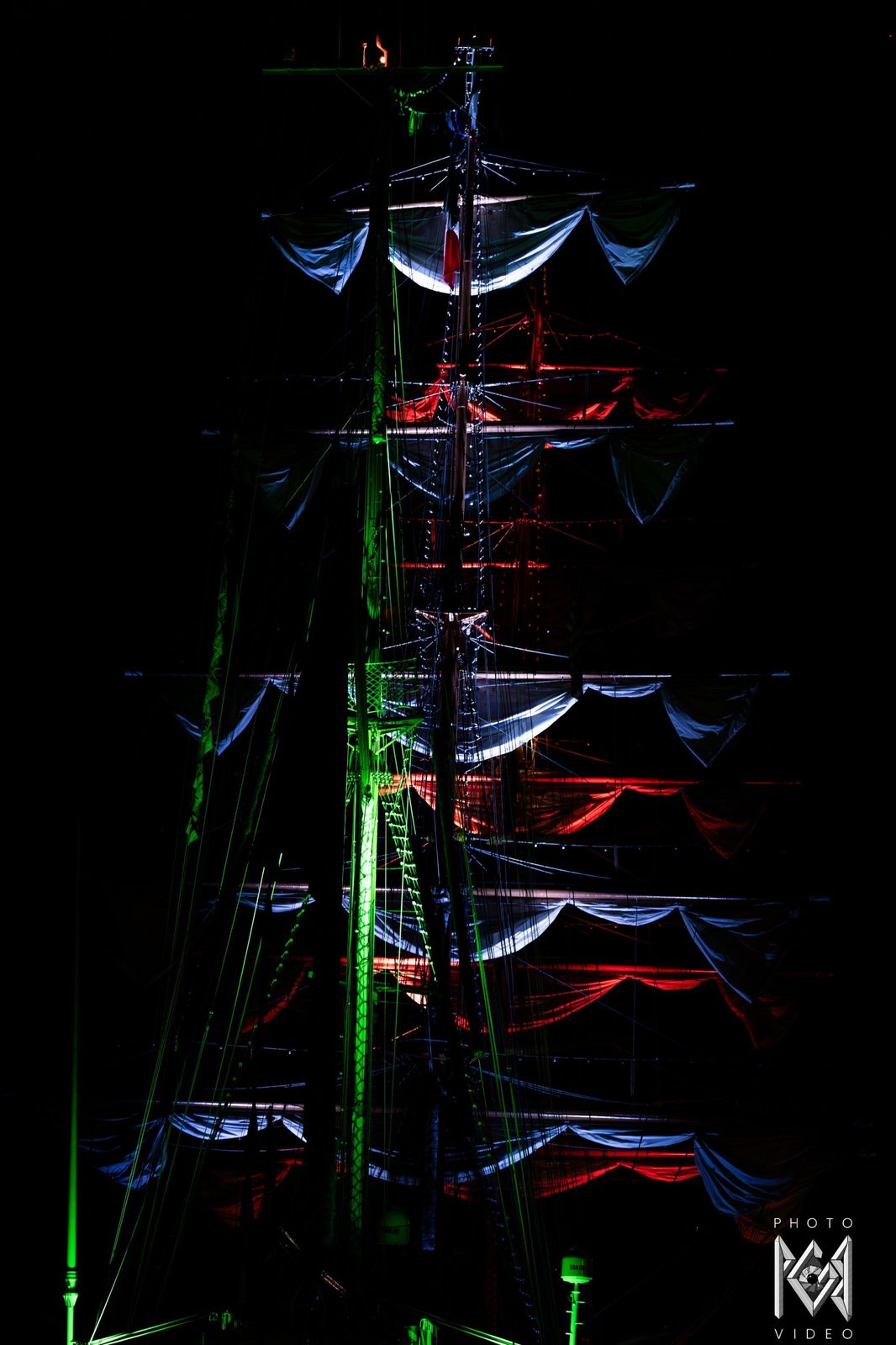 The ship's masts and sails lit in green, white and red against the night sky