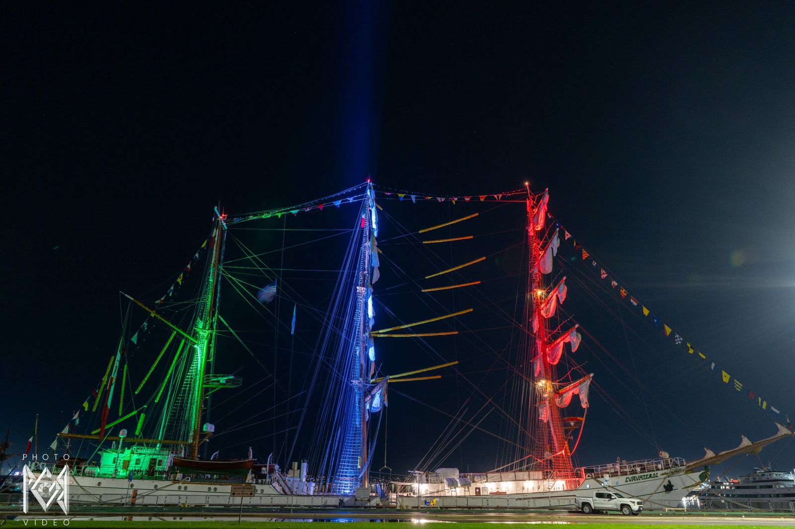 The ship's masts lit in green, white and red across a lawn at night, a beam of light rising behind