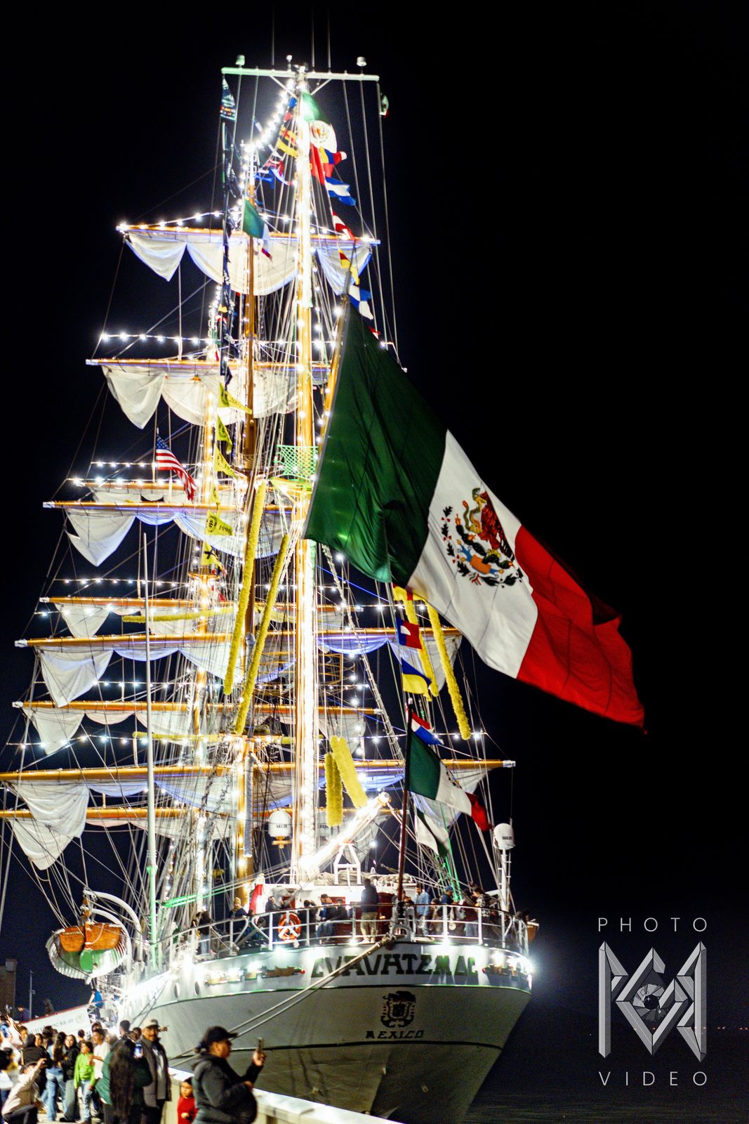 The ship lit from stern to masthead at night with a huge Mexican flag, visitors on the pier