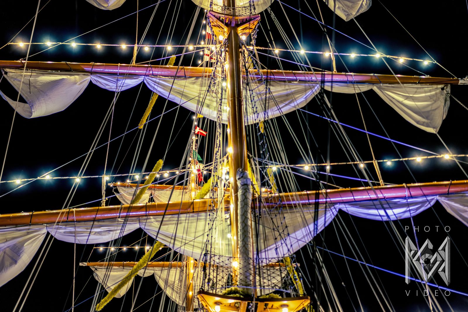 A lit mast with sails and string lights at night