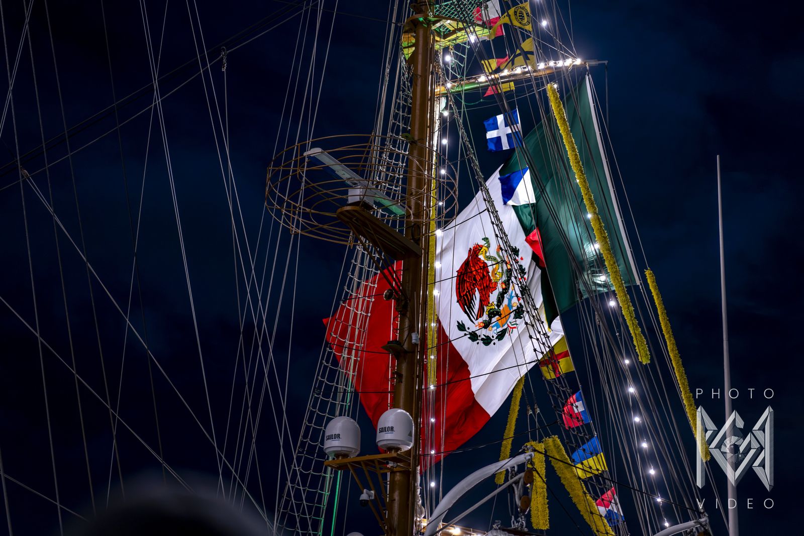The Mexican flag and signal flags beside a mast with a lookout basket at night