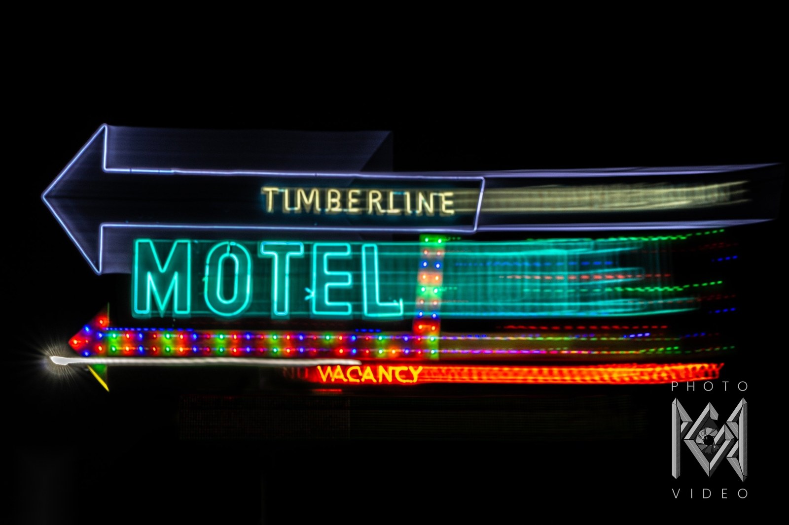 The Timberline Motel neon sign, teal MOTEL lettering and a bulb-lined arrow stretched sideways by a long exposure, VACANCY glowing red below