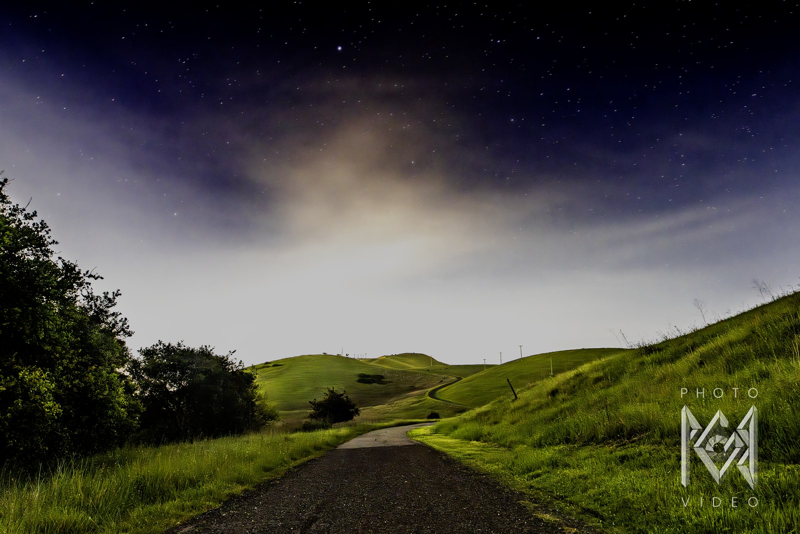A paved path winding up a green hill under a starry night sky, hazy glow along the horizon