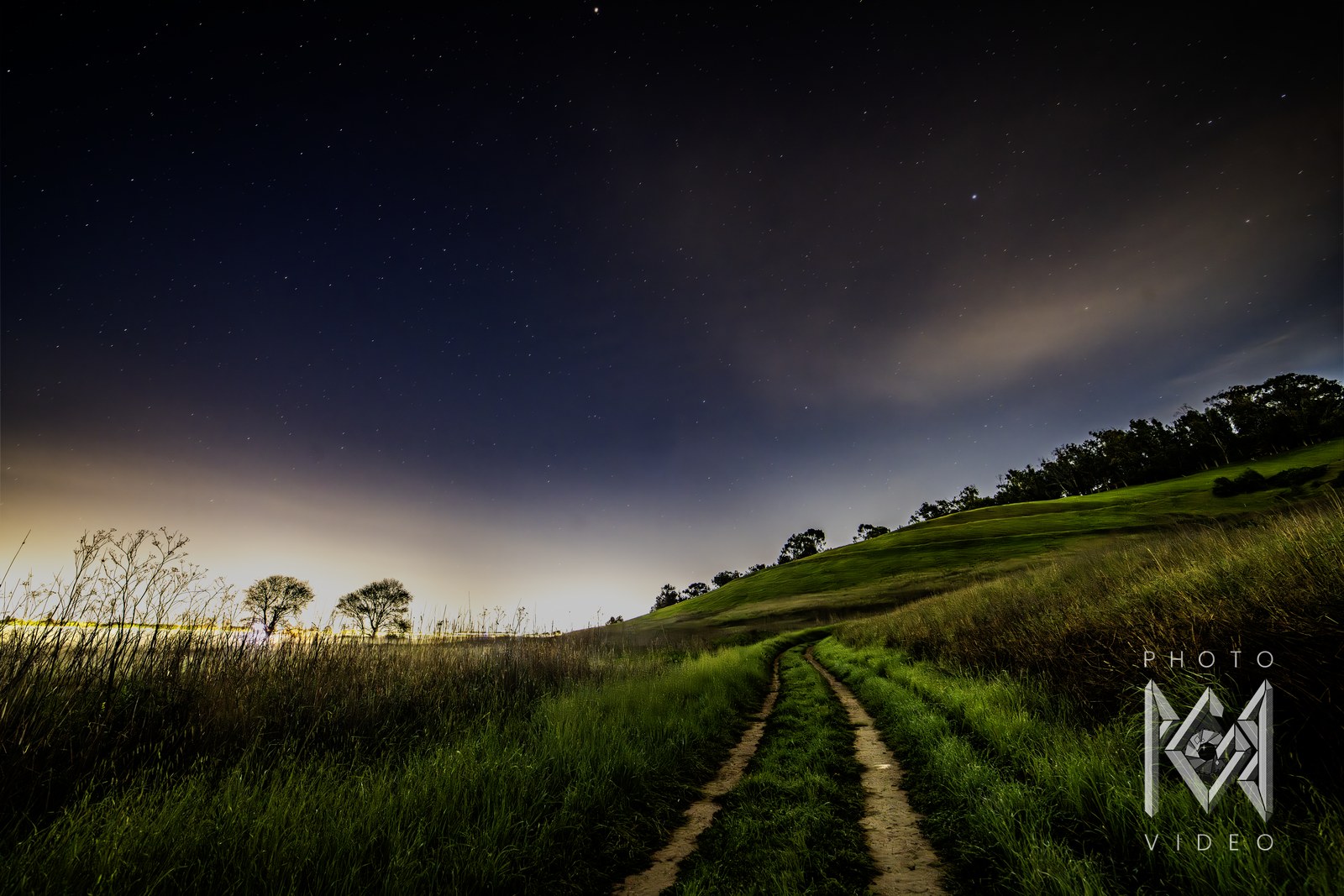 Tractor ruts leading through tall grass toward two trees against a warm dusk glow, green hills beyond