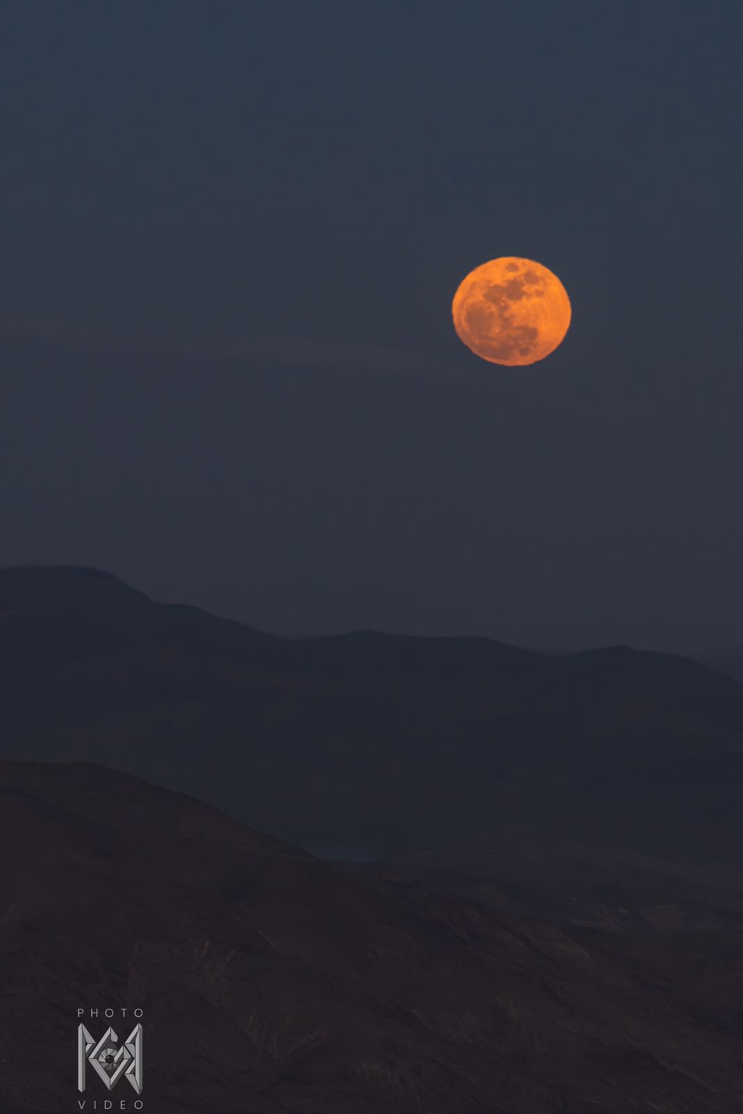 A small orange moon rising over dark, layered desert mountains at dusk